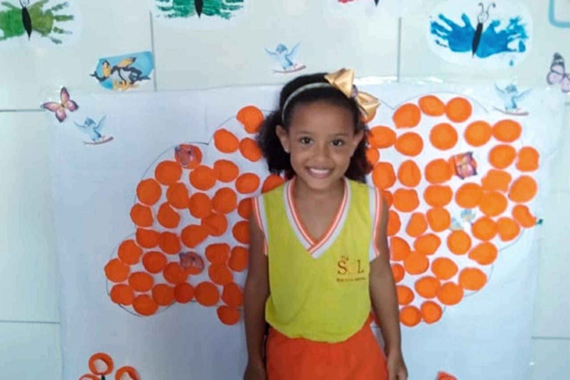 Foto de menina sorrindo em frente á um painel de arte artesanal
